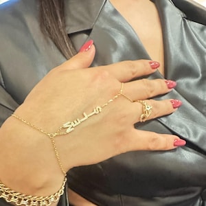 Puede incluir: Una pulsera de mano dorada con la palabra "Suleis" en escritura árabe, adornada con pequeños cristales. También se ve un anillo a juego y una pulsera de cadena dorada. La mano lleva esmalte de uñas rojo.