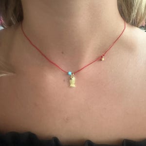 Puede incluir: Un delicado collar de hilo rojo con un pequeño colgante de pez dorado. El collar también incluye una pequeña cuenta turquesa y una cuenta dorada. El collar se lleva alrededor del cuello.