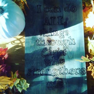 May include: A wooden sign with a blue-grey stain, featuring the text "I can do ALL things through Christ who strengthens me". The sign is surrounded by fall leaves.