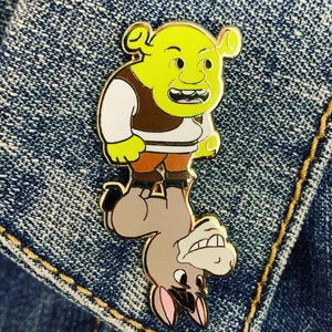 May include: A gold-colored enamel pin featuring Shrek from the Shrek movie franchise riding a donkey. Shrek is green and has a wide grin. The donkey is brown and has a surprised expression.