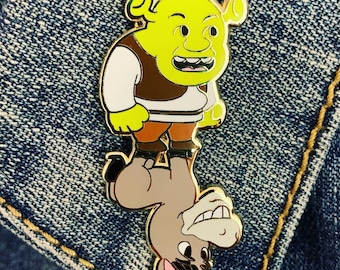 May include: A gold-colored enamel pin featuring Shrek from the Shrek movie franchise riding a donkey. Shrek is green and has a wide grin. The donkey is brown and has a surprised expression.