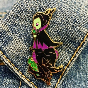 May include: A gold-tone enamel pin featuring a cartoon depiction of Maleficent from Disney's Sleeping Beauty. She is wearing a purple and black dress and has green skin. She is riding a black dragon with green accents.