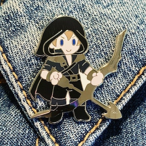 May include: A black and gold enamel pin depicting a cartoon character wearing a black hooded cloak and holding a bow and arrow. The character has blonde hair and blue eyes.