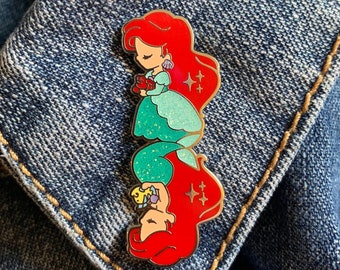 May include: A vertical enamel pin featuring a cartoon mermaid with red hair and a teal dress. The pin has a gold outline and glitter accents. The mermaid is holding a red object and has a pair of earrings. The pin is on a denim background.