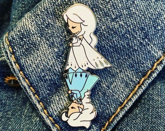 May include: A white and blue enamel pin featuring two cartoon characters. The top character is a girl with long white hair wearing a white dress and blue pants. The bottom character is a girl with long white hair wearing a blue dress. The pin is on a blue denim background.