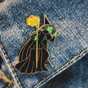 Puede incluir: Una bruja de piel verde con un sombrero puntiagudo y un vestido negro sostiene una escoba amarilla. La bruja lleva un cinturón dorado y tiene la cara verde. La imagen es un pin sobre un fondo de mezclilla.