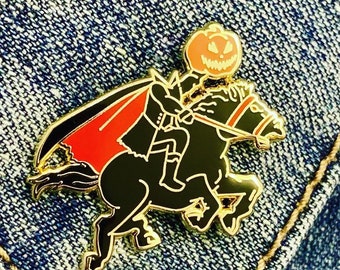 May include: A gold-tone enamel pin featuring a black horse with a red cape and a pumpkin head rider. The horse is galloping and the rider is holding the reins.