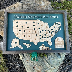 May include: Wooden United States Golf Tour map with a teal background and a gray frame. The map features cutouts for golf courses and includes small wooden markers. The text "UNITED STATES GOLF TOUR" is at the top.