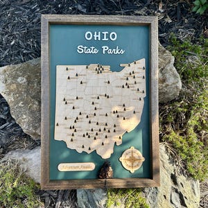 May include: A framed wooden map of Ohio State Parks featuring a teal background, a light wood state outline with engraved park locations marked by small tree icons, a compass rose, and the text "OHIO State Parks" and "Adventure Awaits".
