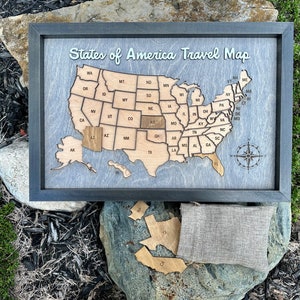 May include: A wooden map of the United States with each state as a separate piece. The map is framed in black and has a compass in the bottom right corner. The states are arranged in a puzzle-like format, and the map is labeled "States of America Travel Map".