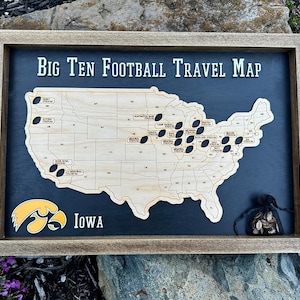 May include: A wooden framed map of the United States with a black background. The map is labeled "Big Ten Football Travel Map" and has a cutout of the United States with black footballs marking locations. The map also features a yellow and black Iowa Hawkeyes logo.