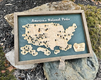 May include: A wooden map of the United States with black tree icons marking the location of each national park. The map is framed in a brown wood frame and has a green background. The text "America's National Parks" is written in a decorative font at the top of the map. The text "National Parks Travel Map" is written in a smaller font at the bottom of the map.