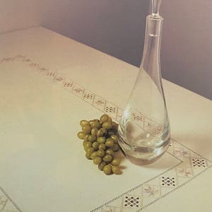 Peut inclure: Un flacon en verre transparent avec un long col et un corps rond posé sur une nappe blanche à motif brodé délicat. Une grappe de raisin vert est placée à côté du flacon.