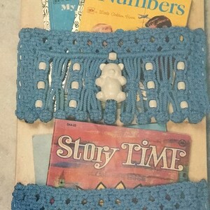 May include: A blue crocheted wall organizer with white beads and a white teddy bear charm. The organizer is holding three children's books: "My First Golden Book of Numbers", "Story Time", and "A Little Golden Book: My First ABC Book".