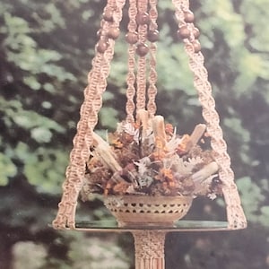 Könnte beinhalten: Ein Makramee-Pflanzenhänger mit einem Holzperlenmuster und einem geflochtenen Korb, der mit getrockneten Blumen gefüllt ist. Der Hänger ist an einem Holzständer befestigt.