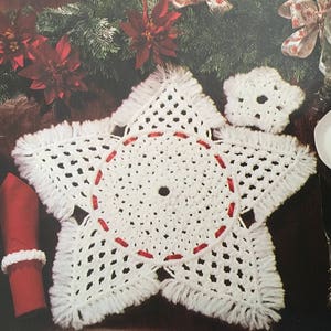 Peut inclure: Une nappe de table en crochet blanc en forme d'étoile avec des points rouges et des franges. La nappe est sur une table brun foncé avec une théière blanche, une tasse et une soucoupe blanches et une serviette rouge.