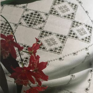 Peut inclure: Une nappe blanche avec un motif brodé délicat en forme de diamant. La nappe est drapée sur une table et présente un arrangement floral d'orchidées rouges au premier plan.