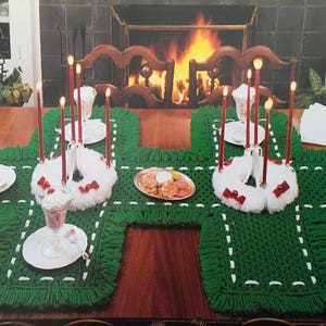May include: A green crocheted table runner with white trim and fringe. The runner is decorated with four white wreaths, each with red bows and candles. There are also four clear glass dishes with red cherries on top.