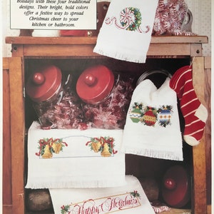 May include: Four white hand towels with embroidered Christmas designs. The towels feature a wreath with a bow, ornaments, bells, and the text "Happy Holidays".