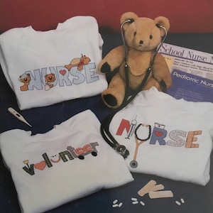 May include: Four white sweatshirts with embroidered designs. The designs include the words "Nurse", "Volunteer", and "Nurse" with a stethoscope and other medical symbols. A brown teddy bear with a stethoscope around its neck is in the background. The text "School Nurse" and "Pediatric Nurse" is visible on a magazine in the background.