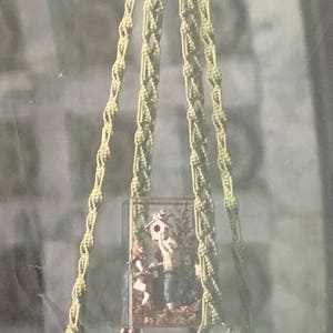 May include: A macrame plant hanger with a shelf, made of natural beige cord. The shelf is holding a small glass box with a miniature scene inside.