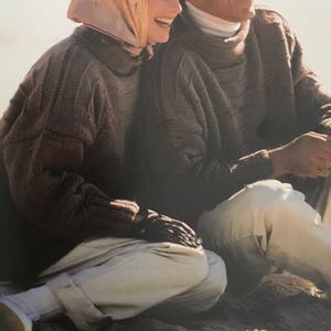 May include: Two people wearing brown knitted sweaters are sitting on a sandy surface. The woman is wearing a white headscarf and the man is wearing a white turtleneck.