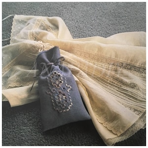 Pode incluir: Uma bolsa de tecido azul com um design bordado branco, amarrada com uma fita marrom, descansando sobre um tecido de renda branco.