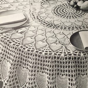 May include: A white, round, crocheted tablecloth with a detailed openwork design. The tablecloth is draped over a table set for a meal, with plates, silverware, and glasses visible. The intricate pattern adds a touch of elegance.