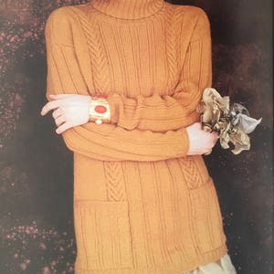 May include: A long-sleeved, mustard yellow sweater with a turtleneck, cable knit design, and two pockets. The sweater is worn by a person with a gold bracelet on their wrist.