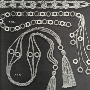 Puede incluir: Tres collares de ganchillo con diferentes diseños. El collar superior es una cadena con nudos, el collar del medio es una cadena con anillos blancos y negros, y el collar inferior es una cadena trenzada con anillos blancos y borlas.