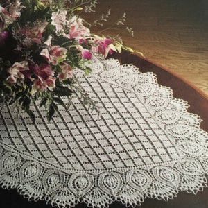 May include: White crocheted table runner with a delicate lace pattern and scalloped edges. The runner is laid on a dark wood table with a bouquet of pink flowers on top.