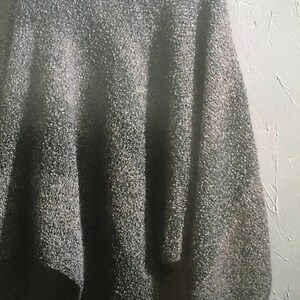 Puede incluir: Un poncho de punto gris con una borla en el borde inferior. El poncho está colgado en una percha de madera contra una pared blanca texturizada.