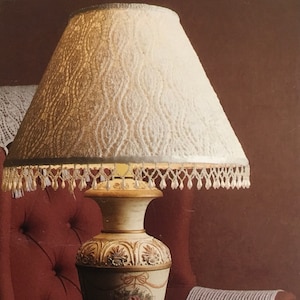 Puede incluir: Una pantalla de lámpara blanca con un delicado patrón de encaje y una franja de cuentas. La pantalla está unida a una base de lámpara pintada de oro con detalles ornamentados.