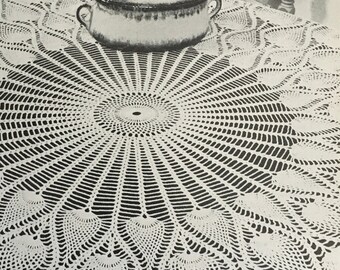 Puede incluir: Un mantel de crochet blanco con un diseño circular que presenta un patrón repetitivo de pequeñas formas puntiagudas. El mantel está colocado sobre una mesa oscura y un plato de cerámica está en el centro.