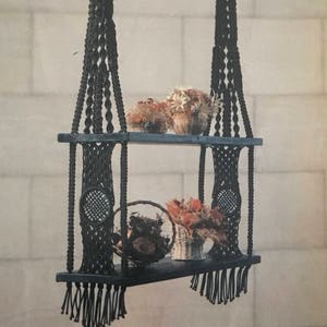 May include: A black macrame hanging shelf with two tiers. The shelf is made of wood and has a black finish. The macrame is made of thick black cord and has a fringe at the bottom. The shelf is hanging from the ceiling and has dried flowers on it.