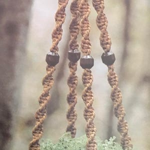 May include: A brown macrame plant hanger with four strands and black wooden beads. The hanger is holding a green plant in a brown pot.