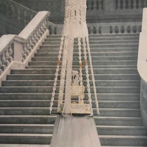 Puede incluir: Un pastel de bodas blanco con un dosel de ganchillo blanco que cuelga de una escalera. El pastel está decorado con flores blancas y una cinta blanca.