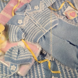 May include: A blue and white crocheted baby outfit with a matching hat. The outfit includes a jacket with a zipper and a pair of pants. The outfit is decorated with white crocheted flowers.