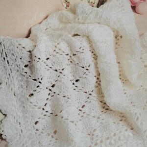 May include: A white crocheted baby blanket with a scalloped edge, resting on a white wicker crib with a pink pillow that says "baby".