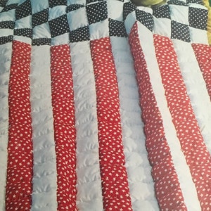 May include: A red, white, and blue quilted blanket with a checkered pattern in the top corner. The blanket is made of fabric with a star pattern and is edged with gold trim.