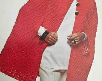 Könnte beinhalten: Ein roter Häkelumhang mit hohem Kragen und drei goldenen Knöpfen. Der Umhang wird über einem weißen Rollkragenpullover und einer weißen Hose getragen.