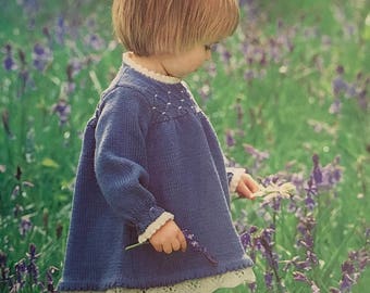 May include: A young child wearing a blue knitted dress with white lace trim stands in a field of purple flowers. The dress has a white collar and a star-shaped design on the front.