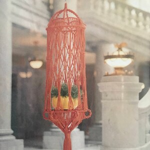 Puede incluir: Un macetero de macramé con un cordón de color terracota y una maceta amarilla con plantas verdes en su interior. El macetero está colgado del techo en una habitación con paredes blancas y un suelo de mármol.