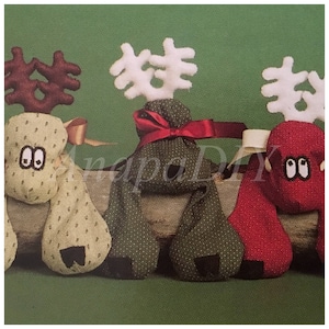May include: Three handmade reindeer ornaments made from fabric. The reindeer are green, red, and beige with brown antlers. The reindeer are sitting on a wooden log.