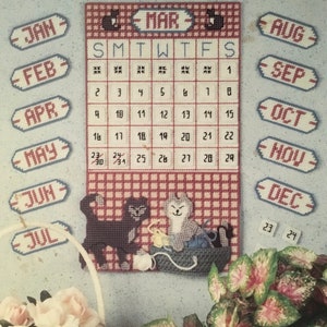 May include: A red and white gingham calendar with a black and white cat and a gray and white cat. The calendar has the months of the year written on it, and the days of the week are abbreviated. The cats are sitting in a basket with yarn.