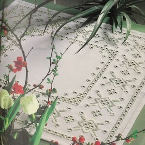 Peut inclure: Un chemin de table blanc brodé avec un motif géométrique. Le chemin de table est sur une nappe verte avec une plante verte en arrière-plan.