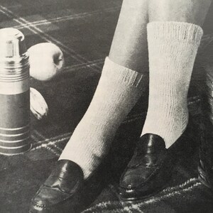 Könnte beinhalten: Ein Paar weiße Stricksocken mit einem schwarzen Leder-Slipper an jedem Fuß. Die Socken sind oben umgeklappt und die Slipper haben ein Quasten-Detail.