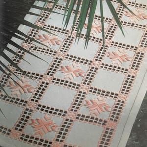 Peut inclure: Un chemin de table blanc avec un délicat motif brodé rose. Le motif présente un motif répétitif de petits carrés avec un motif en forme d'étoile dans chaque carré.