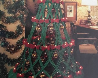 Op de afbeelding: Een groene macramé kerstboom versierd met rode strikken, gouden kerstballen en witte lampjes. De boom heeft een groene tule rok en de tekst "Christmas Tree Delight" is zichtbaar in de afbeelding.