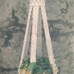 May include: A white macramé plant hanger with a woven design, holding a yellow pot with green leafy plants. The hanger is suspended against a mottled grey and white background, showcasing a bohemian style.